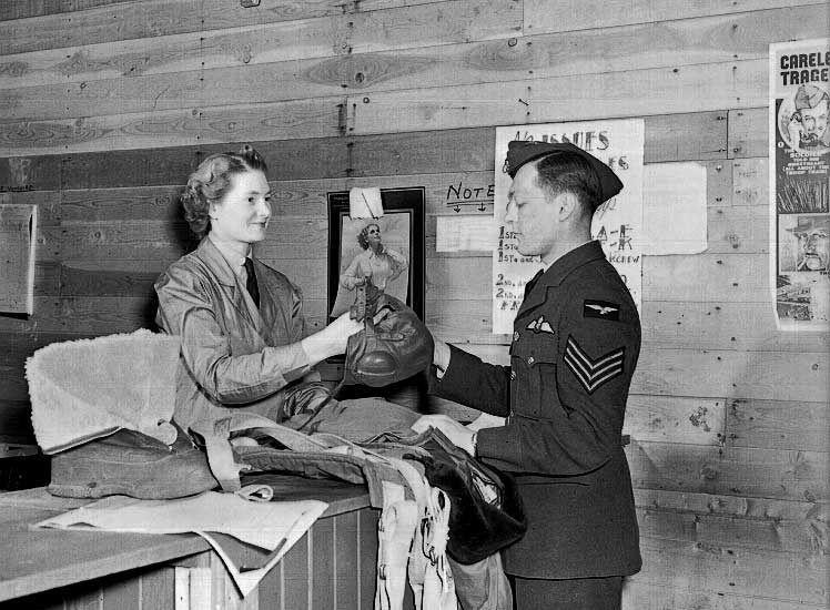 A member of the RCAF Women's Division hands flying gear to an airman in station stores