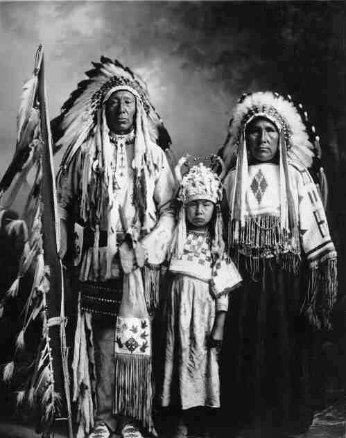 Chief holding eagle feather flagstaff, standing with his wife and child in their beaded clothing and headdresses.
Original caption: