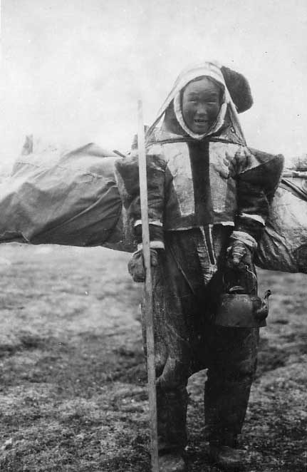 Inuk girl in decorative caribou skin clothing, carrying a stick, a tea kettle and her pack on a burden strap.
Original caption: