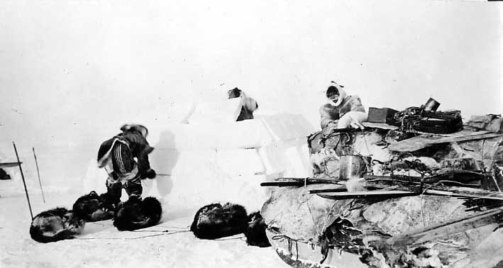 Three Inuit men building an igloo, with packed sled and sleeping Huskies in front.
Original caption:
