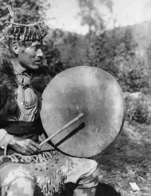 Bella Coola man wearing bear claw headdress and bearskin cape, seated, singing with his round drum.
Original caption: