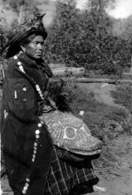 Seated woman wearing button blanket cape and feathered bird skin headdress, holding a large woodcarving on her lap.
Original caption: