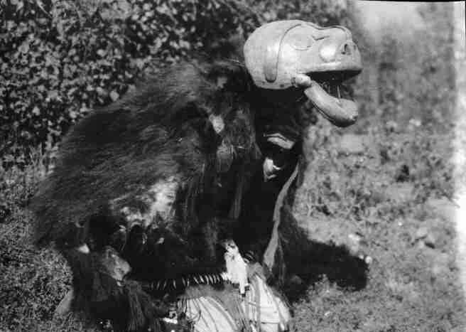 Kneeling dancer hidden under his wooden mask, bearskin cape, and apron decorated with bird beaks and deer hooves.
Original caption: