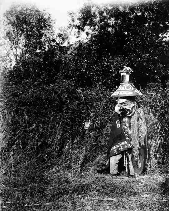 Bella Coola dancer wearing Captain Schooner's two-portion Komokwa hat and mask, and a button blanket robe decorated with cedar fringe over the left shoulder