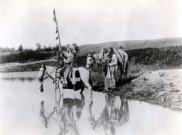 Two men with horses in decorative, beaded hide clothing, at the edge of a river.
Original caption: