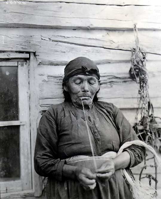 Elderly woman spinning grass outside a log cabin.
Original caption: