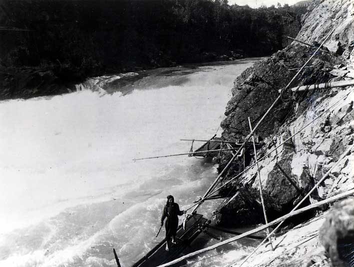 Man holding fish, standing on a platform rigged along a rocky river's edge.
Original caption: