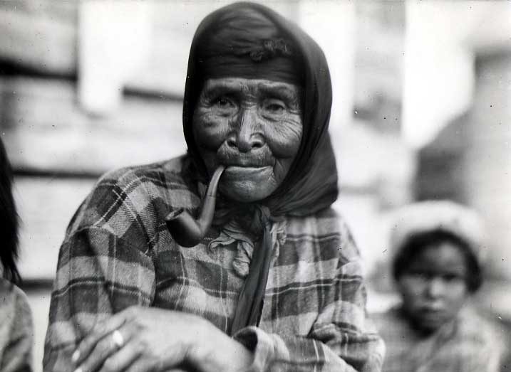 Elderly woman in plaid jacket smoking her pipe, with child in the background.
Original caption: