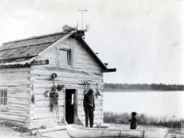 Elderly man outside his log cabin, with child at a canoe