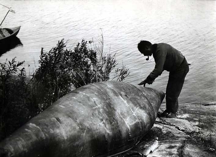 Ojibwa man repairing an upturned birch bark canoe on the shore of Rainy Lake