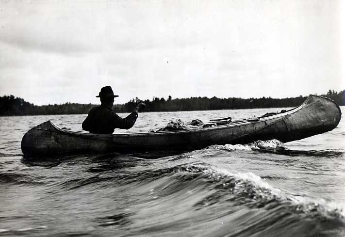 Ojibwa man paddling through waves in loaded birch bark canoe