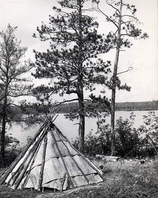 Conical Ojibwa wigwam and campfire, cooking frame at lakeshore.
Original caption: