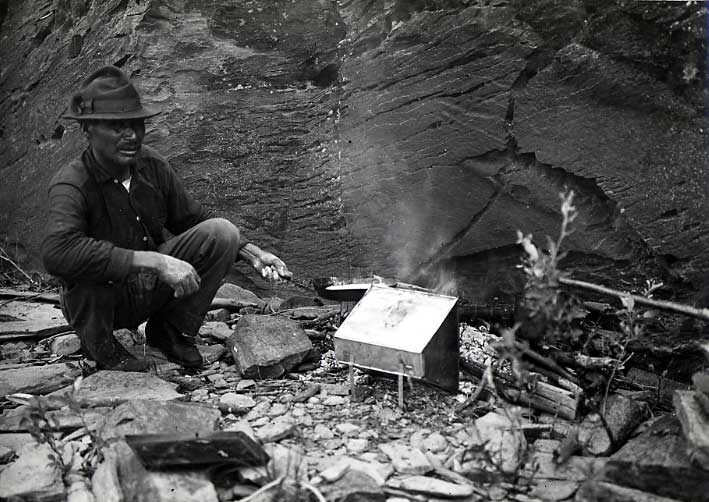 Ojibwa man making bannock over an open fire in front of a rock face