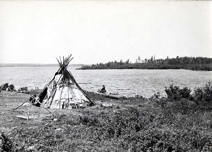Ojibwa birch bark wigwam, campfire tripod and birch bark canoe beside lake.
Original caption: