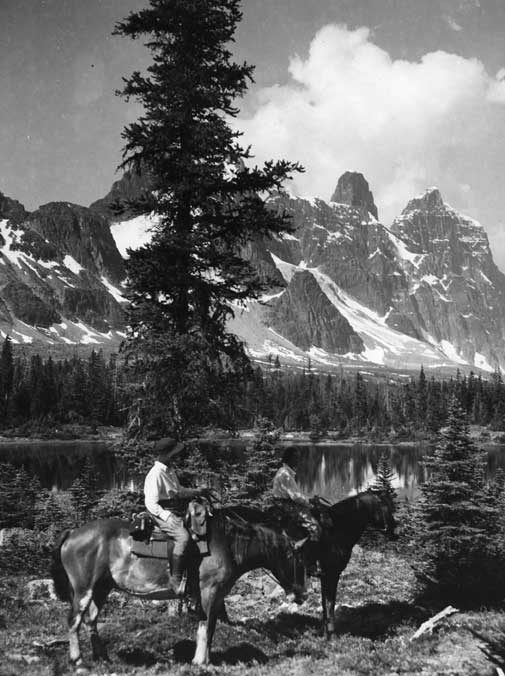 A pair of riders by Moat Lake, with Mts. Turret, Geikie, and Barbican Peaks in the distance