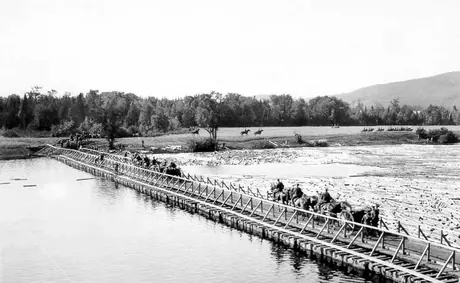 Soldiers on horseback take guns across a bridge to an empty field to train at a WWI training camp