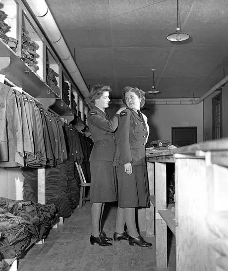 An equipment assistant of the RCAF Women's Division helps an airwoman try on a tunic
