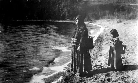 Nakota woman and girl carrying burdens at lakeshore