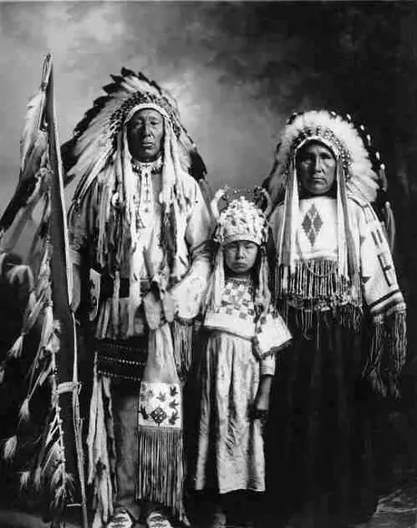 Chief holding eagle feather flagstaff, standing with his wife and child in their beaded clothing and headdresses.
Original caption: