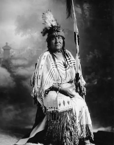 Portrait of White Wings' wife, Blood tribe, wearing a beaded, fringed, hide dress, feathered hat, and carrying an eagle feather staff