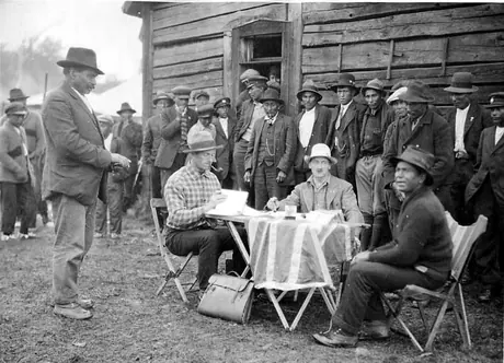 Agents and onlooker seated at table outside a building, dispensing Treaty 3 money to a line-up of Ojibwa men and boys