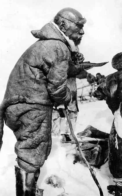 Inuk wearing skin clothing and snow goggles, leaning on a stick, with gun case under his arm. 
Original caption: