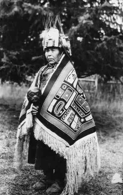 Standing dancer, wearing Chilkat blanket robe, wolf headdress, and carrying a carved wooden face mask rattle.
Original caption: