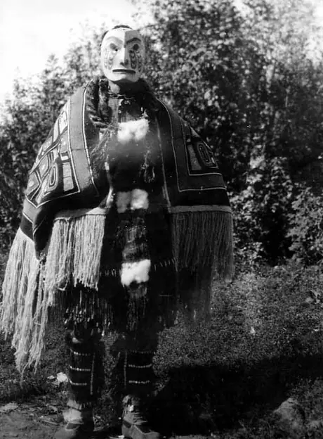 Standing Hamatsa dancer wearing flat wooden face mask, Chilkat blanket robe, cedar rope necklace, and apron with deer hoof rattles on fringes