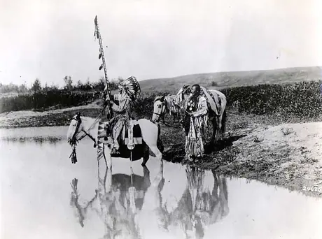 Two men with horses in decorative, beaded hide clothing, at the edge of a river.
Original caption: