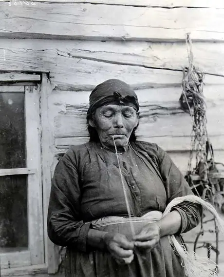 Elderly woman spinning grass outside a log cabin.
Original caption: