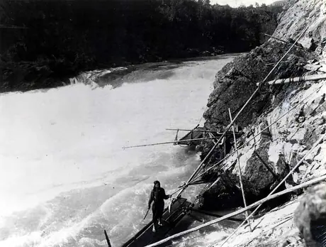Man holding fish, standing on a platform rigged along a rocky river's edge.
Original caption: