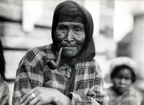 Elderly woman in plaid jacket smoking her pipe, with child in the background.
Original caption: