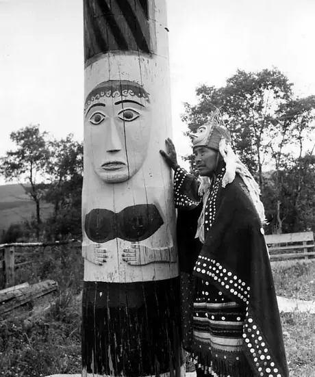 Wet'suwet'en man dressed in button blanket robe, decorative apron, and bird headdress, standing at painted totem pole
