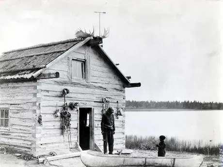 Elderly man outside his log cabin, with child at a canoe