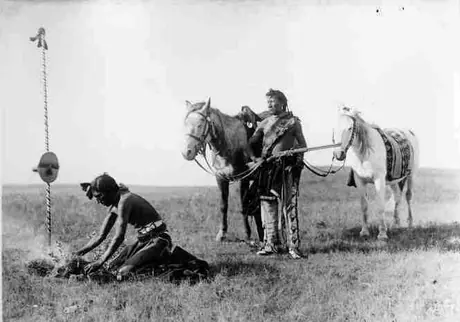 Blackfoot man kneeling at a fire, burning grass, while another man and two horses look on.
Original caption: