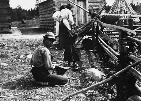 Ojibwa man working at an anvil, with family in background