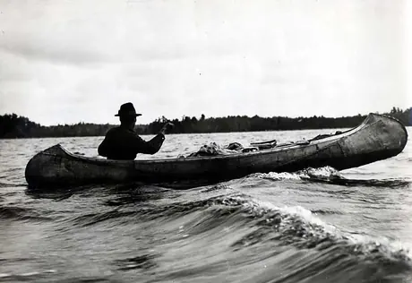 Ojibwa man paddling through waves in loaded birch bark canoe