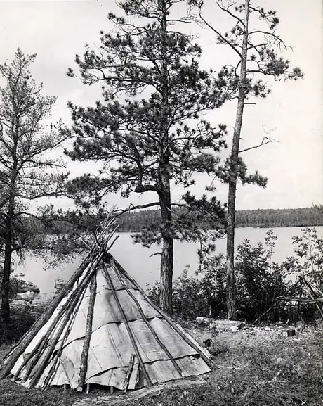 Conical Ojibwa wigwam and campfire, cooking frame at lakeshore.
Original caption: