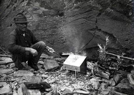 Ojibwa man making bannock over an open fire in front of a rock face