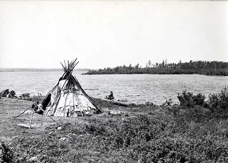 Ojibwa birch bark wigwam, campfire tripod and birch bark canoe beside lake.
Original caption: